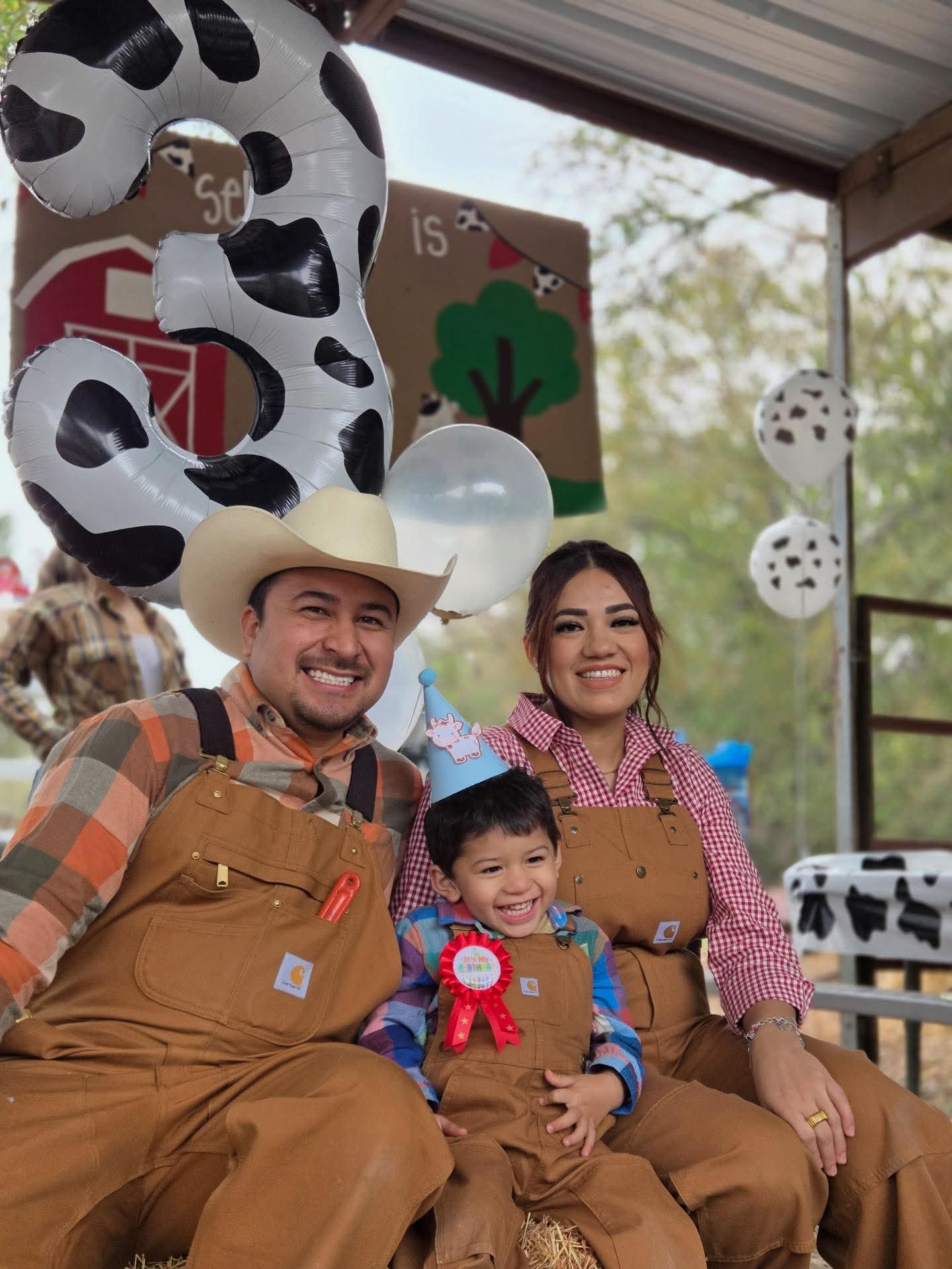Family in farm-themed outfits, posing with a large cow-patterned balloon number 3.