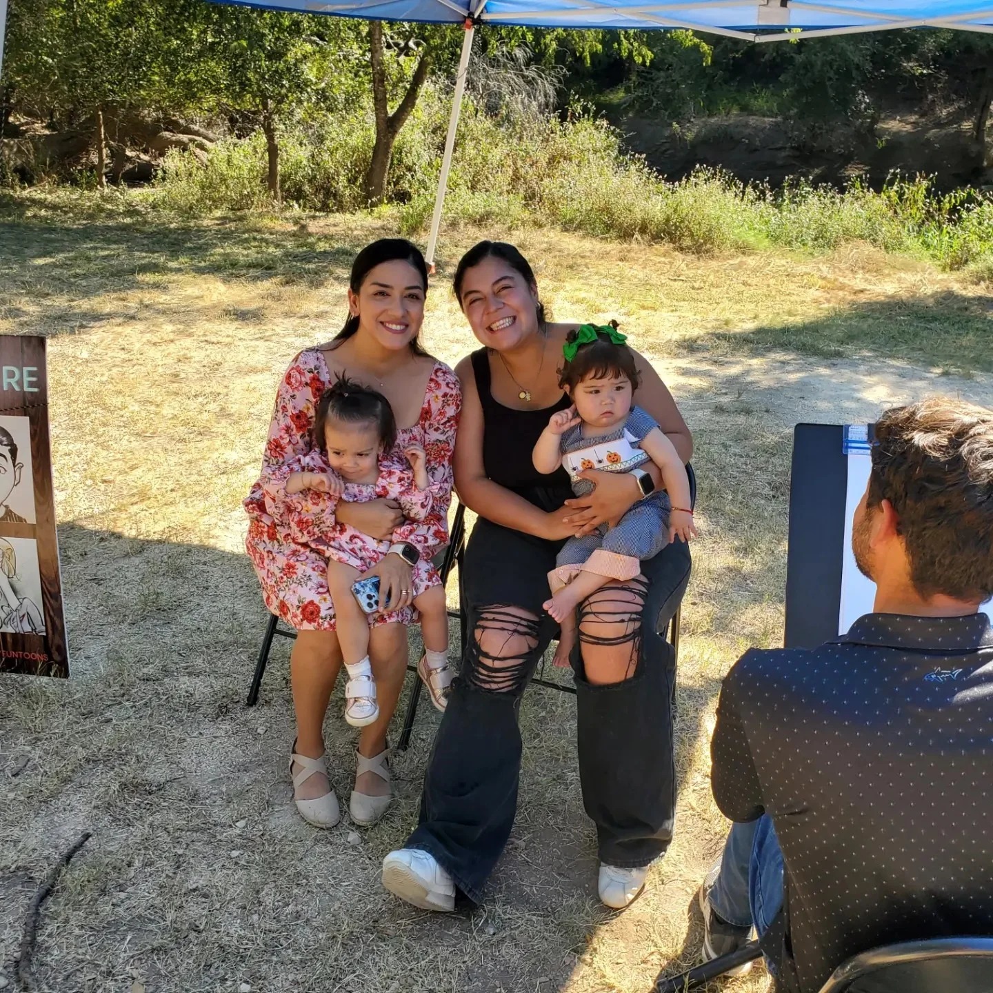 Two women with two toddlers sitting outdoors under a canopy near a portrait artist.