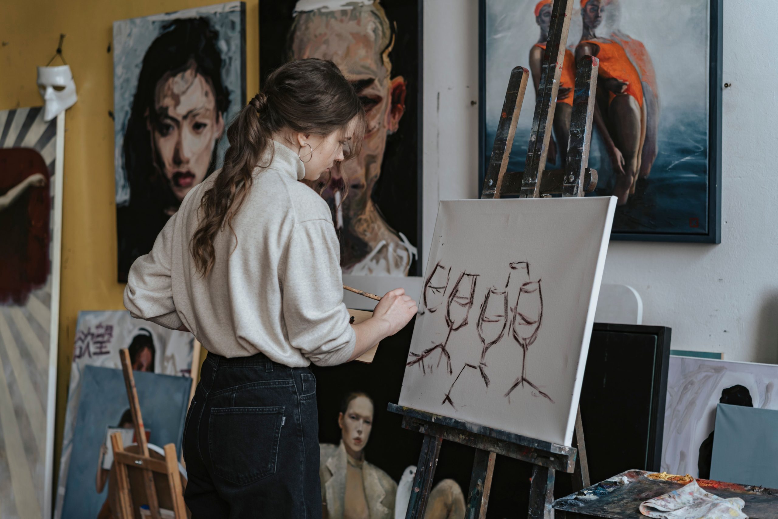 Artist painting wine glasses on canvas in a studio with portraits on walls.